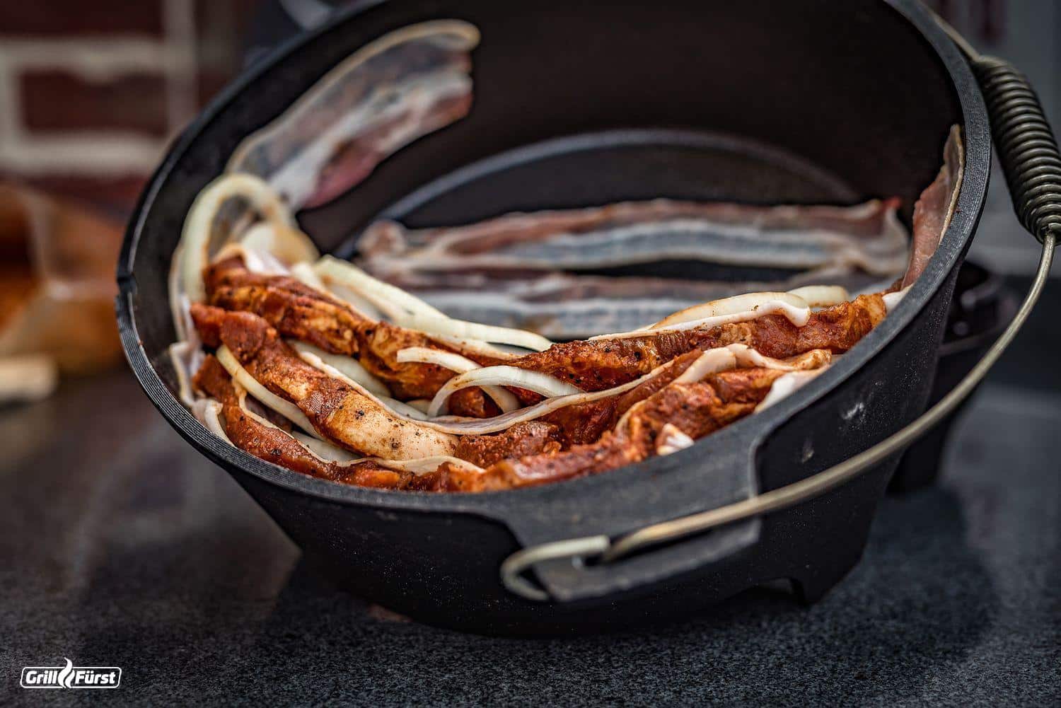 Für die Zubereitung des Schichtfleischs im Dutch Oven werden Bacon, Fleisch, Zwiebeln und Paprika im Topf hergerichtet.