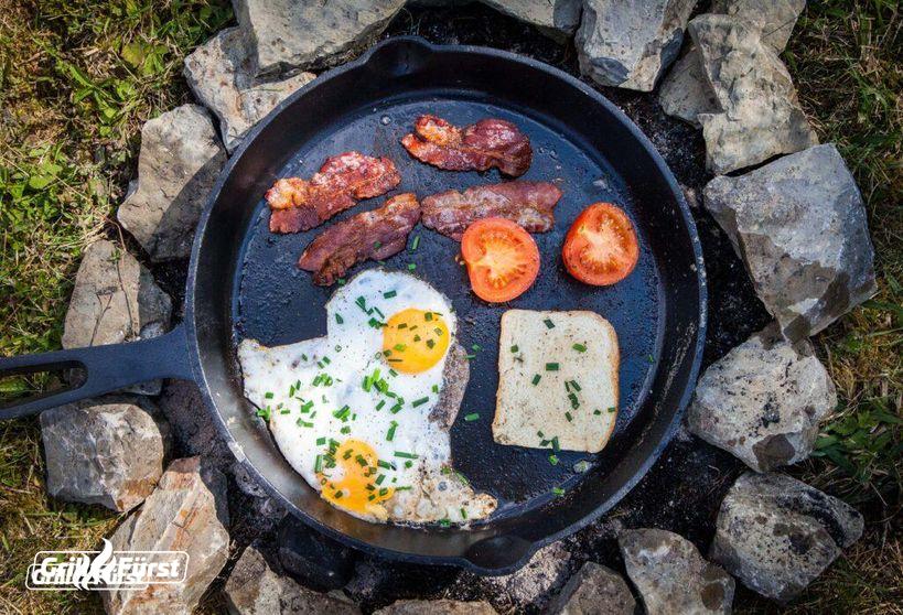 Frühstück mit Bacon, Spiegelei und Toast in Gusseisenpfanne über Feuerstelle zubereitet