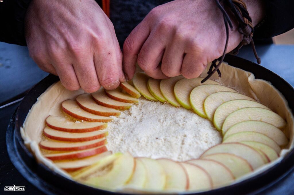 Apfelkuchen wird im Dutch Oven Deckel mit Apfelscheiben belegt.
