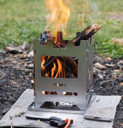 Fennek Premium Hobo Kocher aus Edelstahl mit praktischer Klappkonstruktion und Aschewanne, hier beim Befeuern mit Holz im Outdoor-Einsatz.