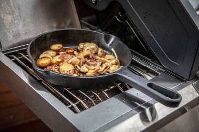 Gusseisenpfanne mit Stiel auf dem Gasgrill mit gebratenen Kartoffeln, Speck und Zwiebeln Gusseisenpfanne mit Stiel auf dem Gasgrill mit gebratenen Kartoffeln, Speck und Zwiebeln
