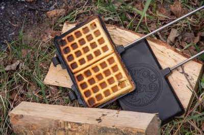 Aufgeklapptes Waffeleisen von Petromax aus Gusseisen mit goldbraun gebackener Waffel auf Holzscheiten im Freien.